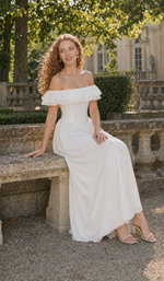 Seated model wearing A Minuet at Versailles white off-shoulder maxi dress in a sunlit Versailles garden.