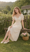Model wearing the Daisies After The Rain Corset Dress in a blooming cottage garden, featuring a romantic floral midi dress with puff sleeves and a smocked waist.