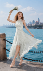 Woman wearing a green halter handkerchief hem chiffon dress holding flowers at a scenic waterfront promenade.