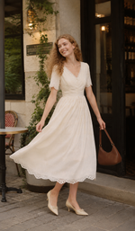 Woman holding a bouquet while wearing The Cream Memory Dress with elegant embroidery and scalloped eyelet hem in a café setting.