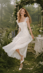 Model smiling in The Rose Oath Dress in white, featuring ruffle straps and a flowing midi silhouette in a dreamy garden.