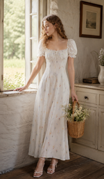 Woman wearing a white cottagecore floral maxi dress in a rustic sunlit room with vintage furniture and countryside views.