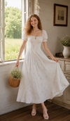 Woman wearing a white floral puff sleeve maxi dress standing beside a sunlit window in a romantic cottage-inspired interior.